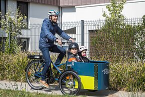 Un homme à vélo portant un casque transporte à l'avant du vélocargo estampillé Vélila 2 enfants. Ils sont en sécurité dans un