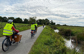 Plusieurs personnes se baladent à vélo sur une piste cycclable avec gilet fluo de sécurité et casqu. UN des vélo est un vélo cargo qui permet de transporter deux enfants à l'avant du vélo en toute sécurité.